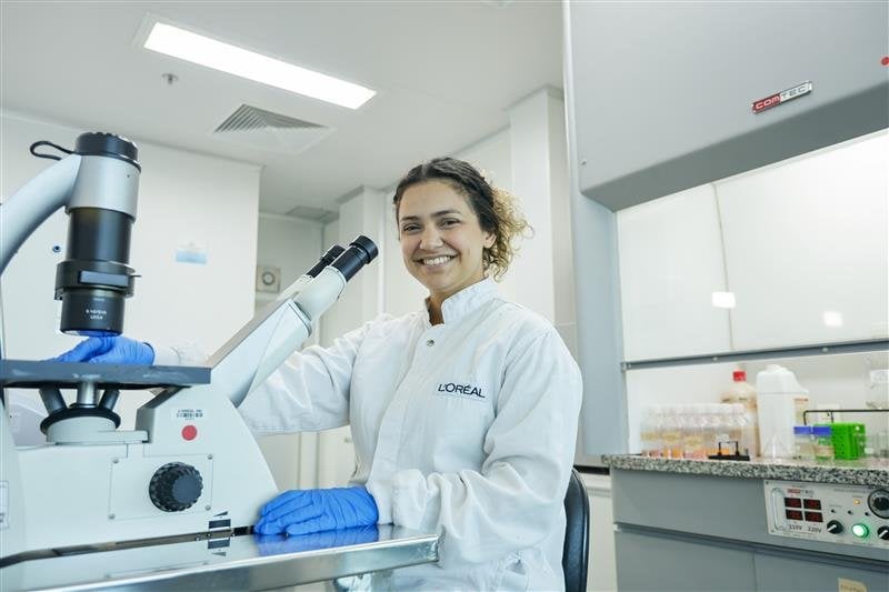 Mulher em um laboratório, sorrindo próxima a um microscópio.