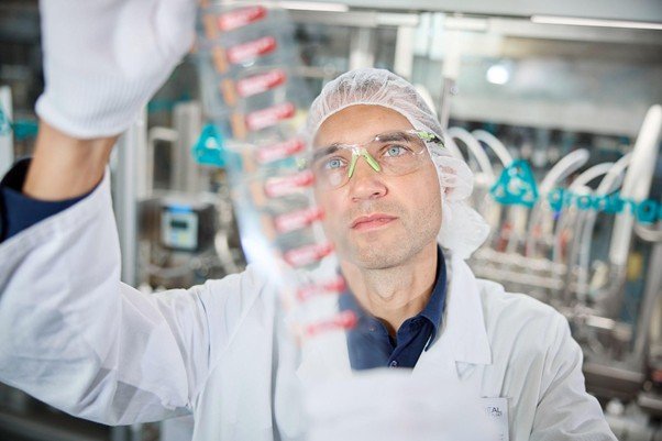 Chemiker in weißem Kittel, Kopfhaube und Schutzbrille schaut Probe an. Im Hintergrund sieht man verschwommen verschiedenen industrielle Gerätschaften.