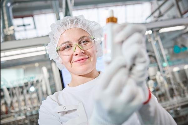 Chemikerin in weißem Kittel, Kopfhaube und Schutzbrille hält ein Flasche mit Pipette. Im Hintergrund sieht man verschwommen verschiedenen industrielle Gerätschaften.