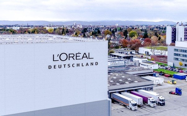 Fabrikgelände Karslruhe mit großem weißem Gebäudeblock mit Aufschrit L'Oréal Deutschland links im Vordergrund. Daneben sieht man ein Gebäude an dem LKWs be- oder entladen werden.