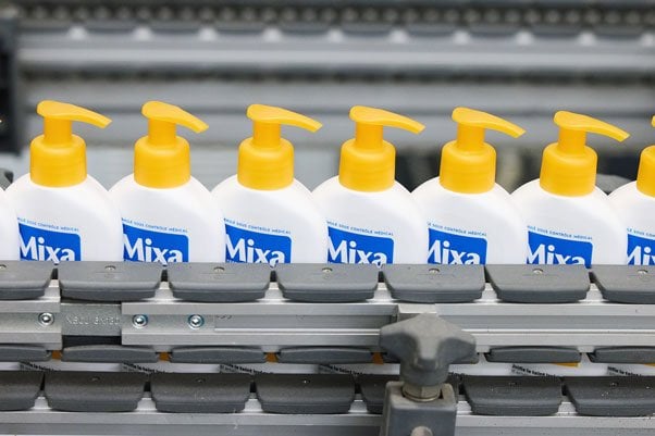 Reihen weißer Mixa-Lotionflaschen mit gelbem Spender, liegen auf einem grauen Förderband.