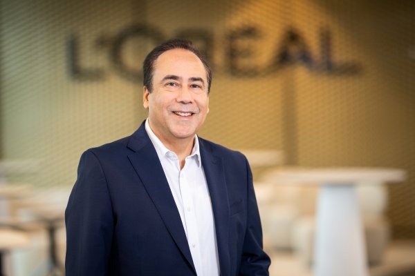 Direktor Loreal Werk Karlsruhe Vishal Sahgal steht vor einem L'Oréal Logo an der Wand. Er trägt ein Hemd ohne Krawatte und eine offene Anzughose