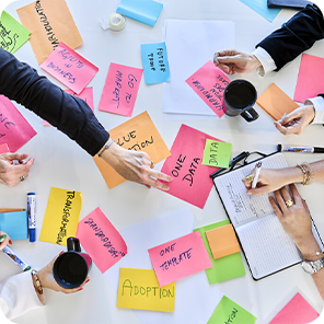 Auf einem Schreibtisch liegen verschiedene bunte Zettel, es sitzen mehrere Personen um den Tisch und arbeiten