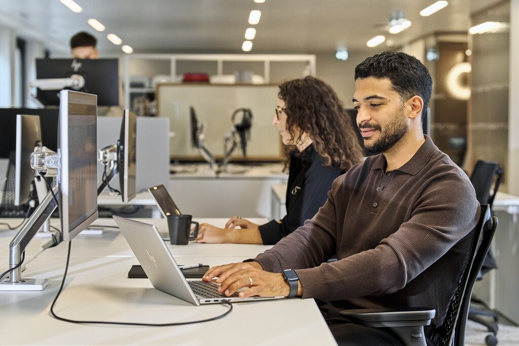 Ein Mann und seine Kollegin im Hintergrund sitzen gemeinsam im Großraumbüro und arbeiten
